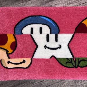 Puede incluir: Una alfombra rosa con una estrella blanca, un hongo azul, un hongo amarillo y una hoja verde. La estrella y los hongos tienen caras sonrientes.