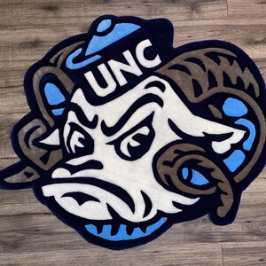 May include: A blue and white rug with a cartoon ram wearing a blue and white hat with the letters "UNC" on it.