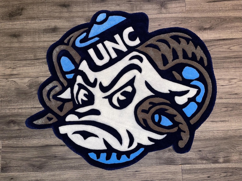 May include: A blue and white rug with a cartoon ram wearing a blue and white hat with the letters "UNC" on it.
