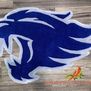 May include: Blue and white rug with a stylized image of a wildcat's head. The rug is handcrafted in Kentucky.