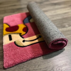 Puede incluir: Una alfombra de dibujos animados de color rosa y amarillo en forma de hongo con un respaldo gris. La alfombra est&aacute; enrollada y el respaldo es visible.
