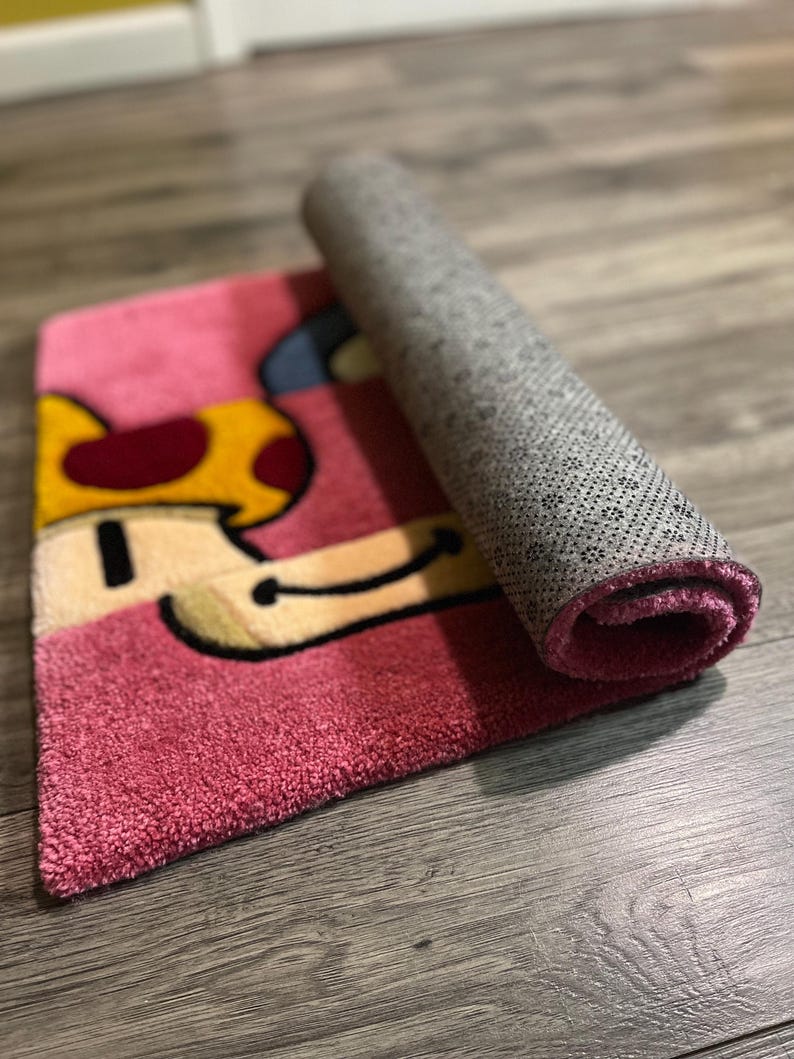 Puede incluir: Una alfombra de dibujos animados de color rosa y amarillo en forma de hongo con un respaldo gris. La alfombra est&aacute; enrollada y el respaldo es visible.