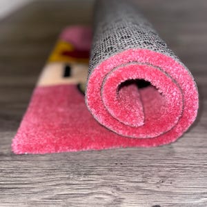 Puede incluir: Una alfombra rosa y gris enrollada con un dise&ntilde;o de dibujos animados. La alfombra tiene un respaldo gris y una pila rosa.