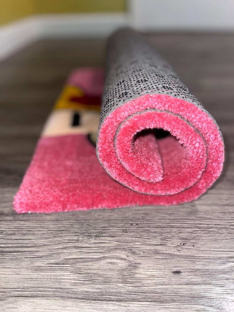 Puede incluir: Una alfombra rosa y gris enrollada con un dise&ntilde;o de dibujos animados. La alfombra tiene un respaldo gris y una pila rosa.