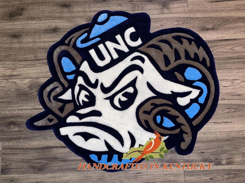 May include: A blue and white rug with a cartoon ram wearing a blue hat with the letters UNC on it. The rug is handcrafted in Kentucky.