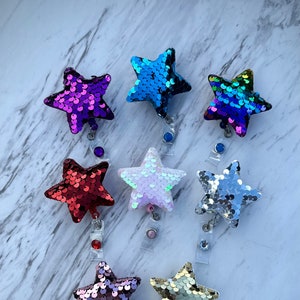 May include: A collection of sequined star-shaped badge reels in various colors, including purple, blue, red, white, silver, and gold. Each star is attached to a clear plastic reel with a clip.