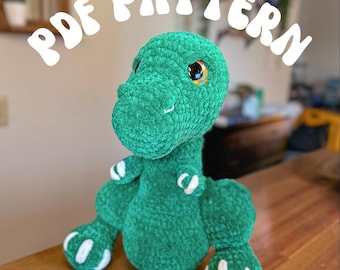 May include: A green crocheted dinosaur toy named Timmy the T-Rex. The toy has white accents on its feet and a yellow eye with black pupils. The image includes the text "PDF PATTERN".