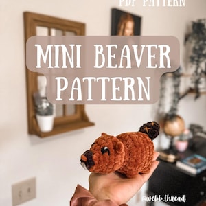May include: A crocheted plush beaver toy in brown and black, held in a hand with a pink hair scrunchie. The text "PDF PATTERN" and "MINI BEAVER PATTERN" is visible in the background. The image is captioned "@webb_thread".