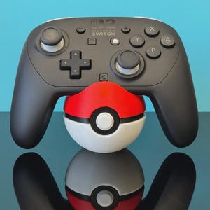 Op de afbeelding: Een zwarte Nintendo Switch Pro-controller die rust op een rood-witte bolvormige standaard die lijkt op een Poké Ball. De controller is voorzien van analoge sticks, een richtingspad en knoppen met de letters X, Y, A en B.