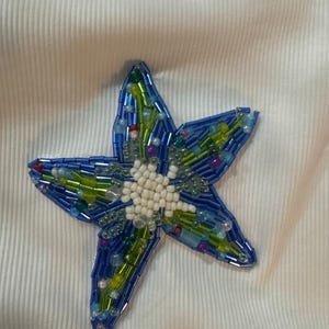 May include: A white ribbed fabric garment featuring a five-pointed star appliqué. The star is intricately decorated with a mix of blue and green glass beads, with a cluster of small white beads forming a circular center point on the star.