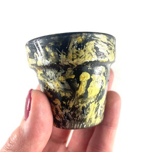 May include: A small black terracotta pot with gold and silver paint splatters. The pot is held in a hand.