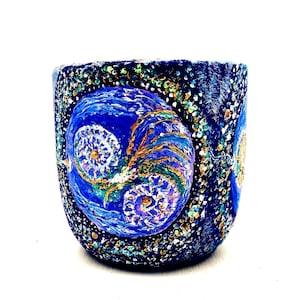 Peut inclure: Pot en céramique cylindrique peint à la main, avec un fond bleu foncé. Le pot présente un motif abstrait tourbillonnant dans des tons de bleu, d'or et de violet. L'extérieur est texturé avec de petits ornements colorés.