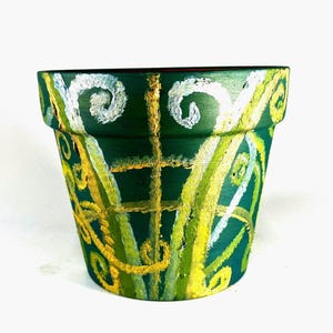 Puede incluir: Una maceta de cerámica verde oscuro pintada a mano. La maceta está decorada con espirales blancas y líneas verticales y horizontales amarillas y verde claro. La maceta está vacía y lista para plantar.