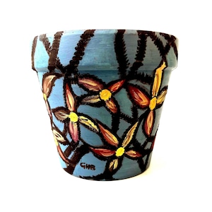 Könnte beinhalten: Handbemalter Keramik-Blumentopf. Der Topf ist hellblau mit einem dunkelbraunen Netzwerkdesign. Die Blumen sind gelb, rot und braun. Die Initialen "GMB" sind auf die Seite des Topfes gemalt.