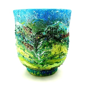 May include: A textured, hand-painted ceramic vase with a landscape design. The vase features a vibrant colour palette of blues, greens, and yellows, depicting a mountain scene. The base is approximately 10 cm in diameter.