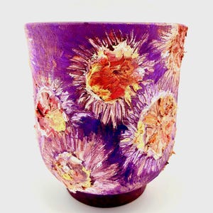 Puede incluir: Un jarrón de cerámica pintado a mano con una superficie texturizada. El jarrón es principalmente morado con diseños florales en relieve en tonos de rojo, naranja y amarillo. La base es de un tono morado más oscuro.