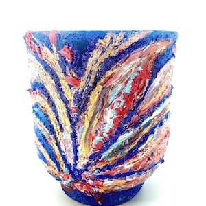 May include: A hand-painted ceramic vase with a textured surface. The vase features an abstract design with strokes of blue, red, yellow, and pink paint. The base and rim are a solid dark blue.