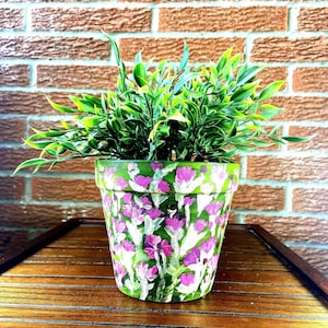 Op de afbeelding: Een handbeschilderde keramische bloempot met een groene basis en een bloemenontwerp in roze en witte tinten. De pot is gevuld met levendig groen blad. De pot staat op een houten oppervlak tegen een bakstenen muur.