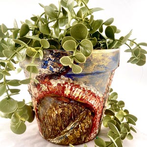 May include: A hand-painted terracotta flower pot with an abstract design in red, blue, and gold. The pot is filled with green, leafy faux eucalyptus. The pot's rim has a gold finish, creating a rustic and artistic appearance.