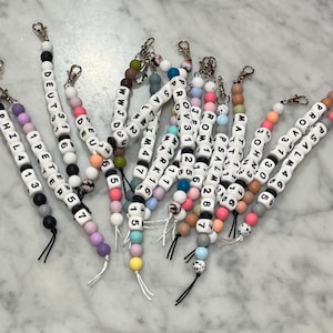 Puede incluir: Una colección de pulseras de cuentas de colores con versículos bíblicos y números de capítulo. Cada pulsera tiene una cuerda negra unida a un cierre plateado.