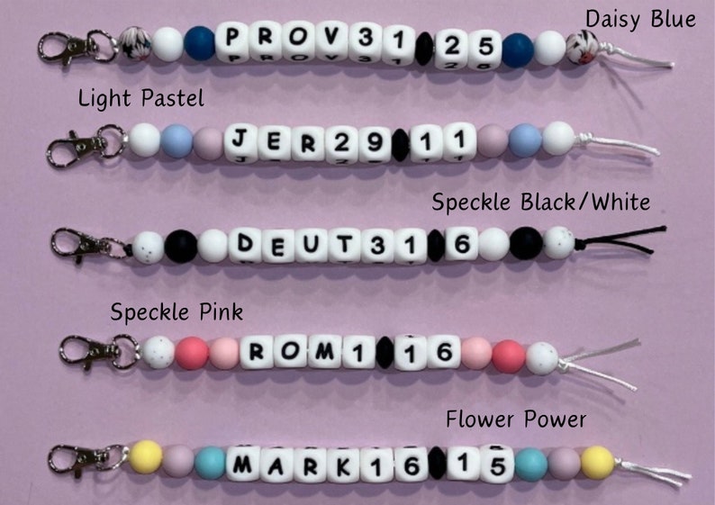 May include: Five colorful silicone bead keychains with different names and numbers. The keychains are in the following colors: daisy blue, light pastel, speckle black/white, speckle pink, and flower power.