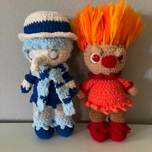May include: Two crocheted plush toys. The first is a blue and white character with a white hat and a long nose. The second is a red and orange character with a large orange hairpiece and a red nose.