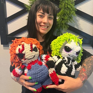 May include: Two crocheted plush toys, one red and blue with a stitched-on face and the other black and white with green hair. The toys are being held by a person with a tattoo on their arm.