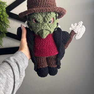 Pode incluir: Boneca de crochê feita à mão de um monstro com rosto verde, chapéu marrom e casaco preto. A boneca tem olhos vermelhos e uma camisa vermelha. Ela está segurando um machado branco e marrom. É um item artesanal único.