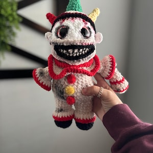 May include: Crocheted clown doll with a white and gray body, red and black accents, and a menacing grin. The doll has a green and yellow cone-shaped hat and large, reflective eyes. A unique handmade item.