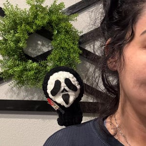 May include: A black and white crochet Ghostface doll holding a small red and silver knife. The doll is perched on a person's shoulder. A green wreath hangs in the background.