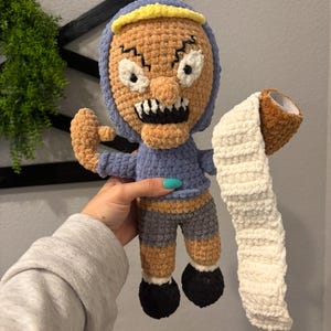 May include: A crocheted plush toy of a cartoon character with a grumpy expression. The toy is wearing a blue hooded shirt, brown pants, and black shoes. The toy is holding a crocheted roll of toilet paper.