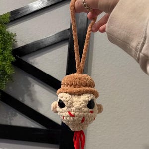 May include: A handmade crochet ornament featuring a stylized face with a brown hat and a red, dangling element. The face is cream-colored with black eyes and red stitching details. The ornament is held by a brown loop.
