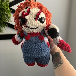 Könnte beinhalten: Eine gehäkelte Puppe mit rot-weiß-blauer Kleidung. Die Puppe hat ein gesticktes Gesicht mit schwarzen Augen und einem gestickten Mund. Die Puppe hält ein schwarzes Mikrofon.