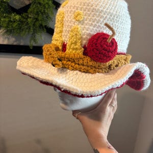 Pu&ograve; includere: Un cappello all'uncinetto fatto a mano a forma di torta. Il cappello presenta una corona bianca con dettagli gialli e marroni, una ciliegia rossa e un'ampia tesa con bordi rossi. Il cappello &egrave; progettato per assomigliare a una fetta di torta.