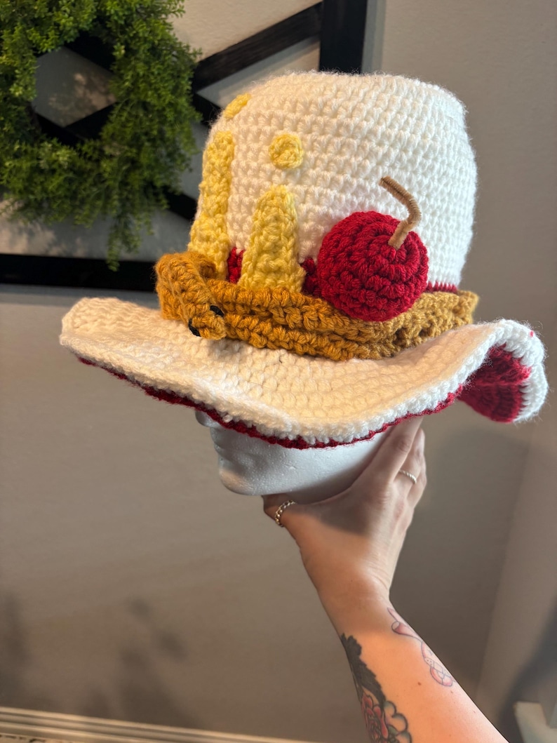 Pu&ograve; includere: Un cappello all'uncinetto fatto a mano a forma di torta. Il cappello presenta una corona bianca con dettagli gialli e marroni, una ciliegia rossa e un'ampia tesa con bordi rossi. Il cappello &egrave; progettato per assomigliare a una fetta di torta.