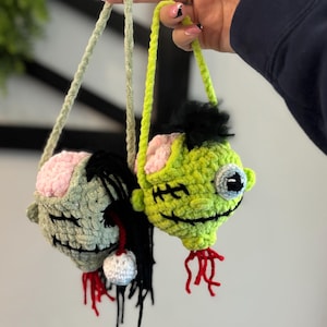 May include: Two crocheted zombie heads with green and pink skin, black hair, and red yarn for blood. The heads are hanging from green and white yarn strings.