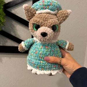 May include: A crocheted plush toy wolf wearing a blue and white floral dress and a matching hat. The wolf has large green eyes and a pink nose.