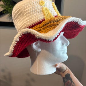 Pu&ograve; includere: Cappello all'uncinetto fatto a mano con tesa larga. Il cappello presenta una corona bianca con accenti gialli, una fascia marrone e una tesa rossa e bianca. Il cappello &egrave; esposto su una testa di manichino bianca.