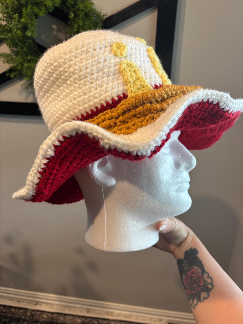 Pu&ograve; includere: Cappello all'uncinetto fatto a mano con tesa larga. Il cappello presenta una corona bianca con accenti gialli, una fascia marrone e una tesa rossa e bianca. Il cappello &egrave; esposto su una testa di manichino bianca.