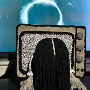 Könnte beinhalten: Handgefertigter Häkel-Fernseher mit einer Figur mit langen schwarzen Haaren, die aus dem Bildschirm auftaucht. Der Fernseher hat ein schwarz-, grau- und beigefarbenes Farbschema mit grauen Pompon-Akzenten. Die Hände sind grau.