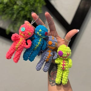 May include: Four colorful crochet voodoo doll keychains. The dolls are pink, blue, purple, and green, each with contrasting stitching and button eyes. They are attached to silver keychains.