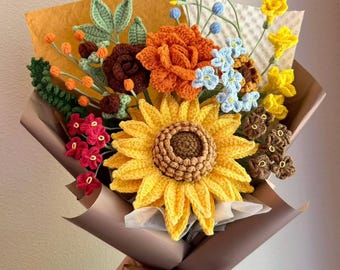 May include: A crochet flower bouquet featuring a large yellow sunflower, orange and brown roses, small red, blue, and yellow blossoms, and green leaves. The arrangement is wrapped in brown paper with a brown ribbon. Text reads luxury Caramel.