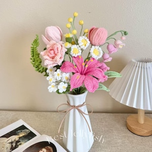 May include: A handcrafted bouquet of crochet flowers in a white vase. The arrangement includes pink lilies, roses, tulips, and daisies, with green leaves and stems. The vase is tied with a light brown ribbon. The overall theme is a decorative floral arrangement.