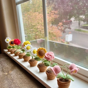 May include: A row of seven crocheted flower pots with different colored flowers. The flowers are pink, yellow, white, red, and orange. The pots are brown and the flowers are sitting on a white windowsill.