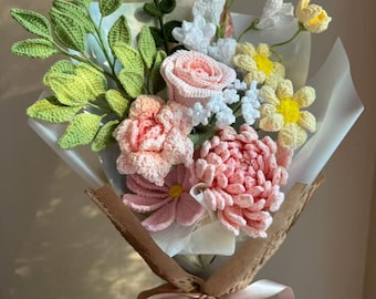 May include: A handmade crochet flower bouquet featuring pink roses, a dense pink dahlia-like bloom, light yellow daisies, and white filler flowers. The arrangement includes textured green leaves and stems, all wrapped in white paper and brown honeycomb paper with a light pink ribbon. The text BestJellyfish is visible.