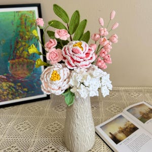 May include: A crocheted flower bouquet in a white ceramic vase. The bouquet includes pink and white flowers with green leaves. The vase has a textured surface.