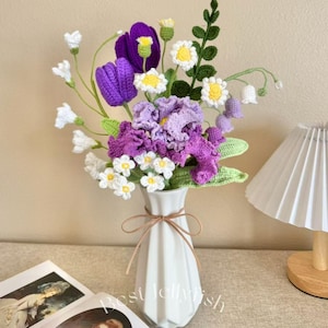 May include: A handmade crochet bouquet of purple tulips, daisies, and other flowers in a white vase tied with a brown ribbon. The flowers are arranged in a decorative display. The vase is on a table next to a book and a lamp.