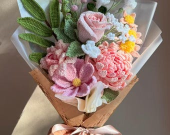 May include: A handcrafted crochet bouquet featuring pink roses, daisies, and other flowers, wrapped in brown paper and tied with a pink ribbon. The bouquet includes green leaves and small yellow flowers, creating a colorful and textured floral arrangement.
