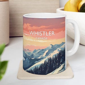 May include: White ceramic mug featuring a scenic mountain landscape with the text "WHISTLER CANADA". The design showcases a sunset sky over snow-capped mountains and evergreen trees. The mug sits on a beige coaster.