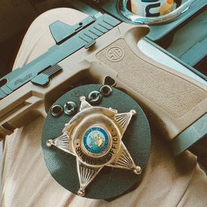 May include: A tan and black handgun with a red dot sight and the Sig Sauer logo. A gold and blue sheriff's badge is below the gun, along with several black rings. The badge is star-shaped and has the words "SHERIFF'S OFFICE" on it.